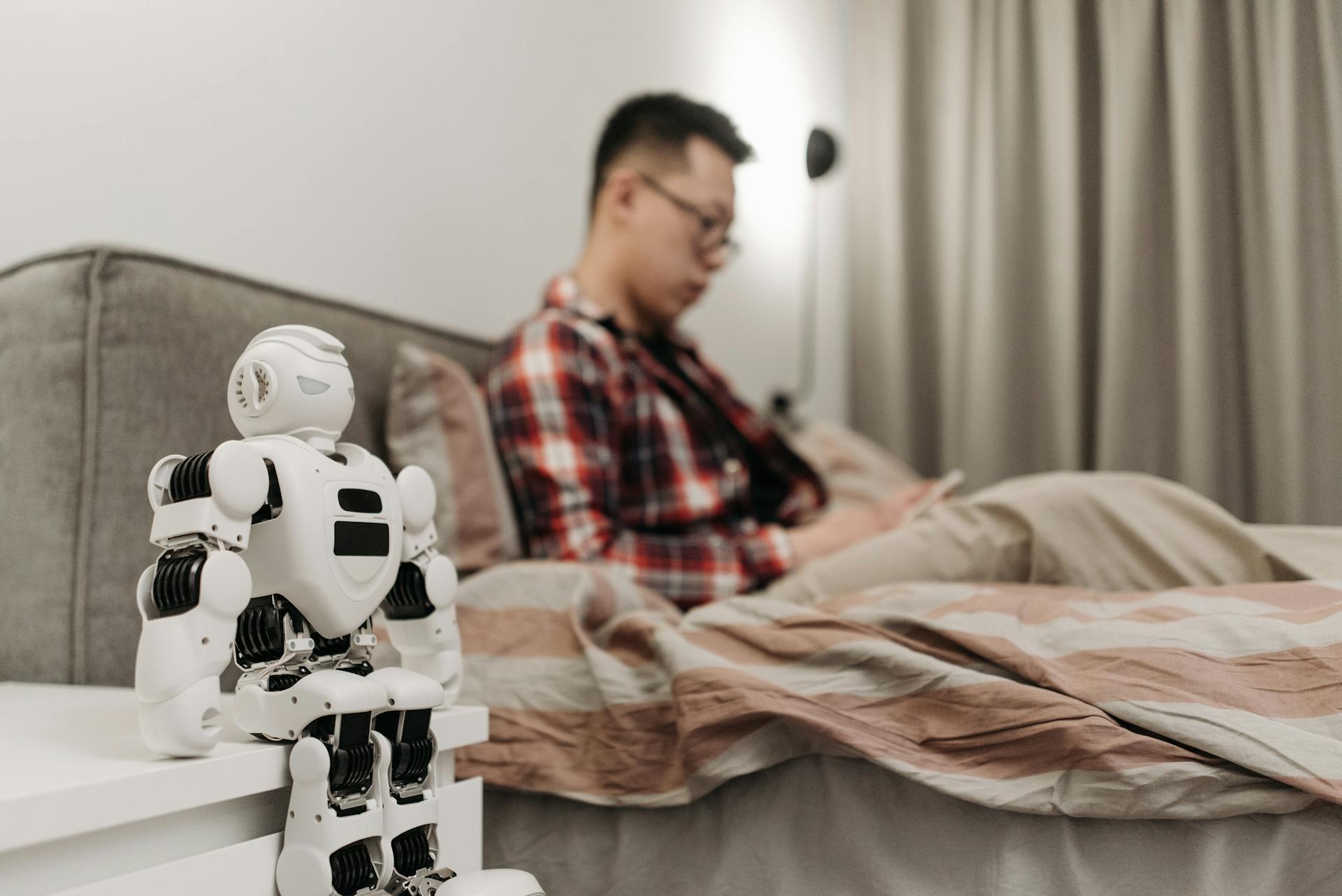 A modern bedroom featuring a robot on a bedside table and a man sitting on a bed.