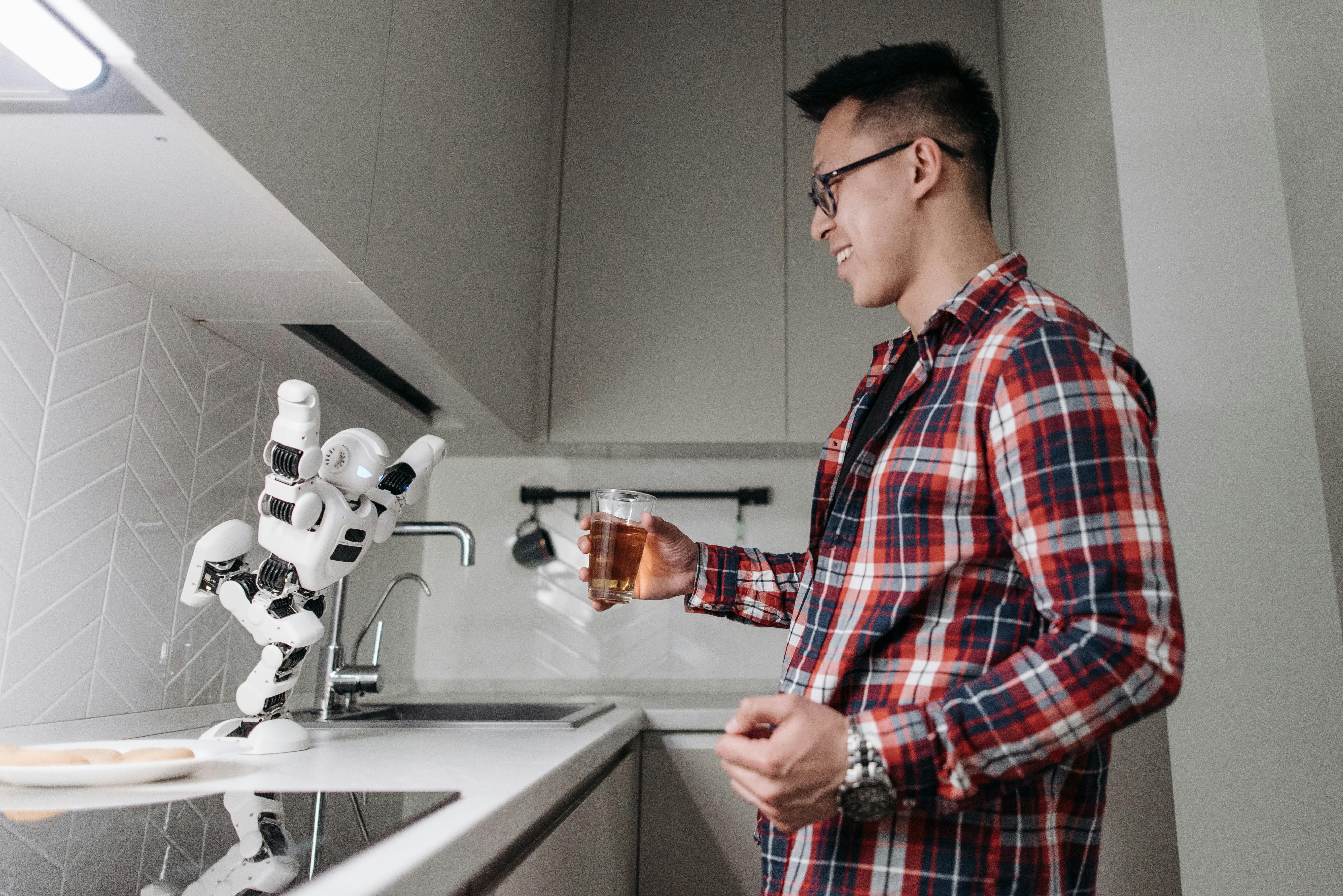 man smiling in plaid long sleeve shirt holding a glass while playing on a dancing robot on kitchen counter