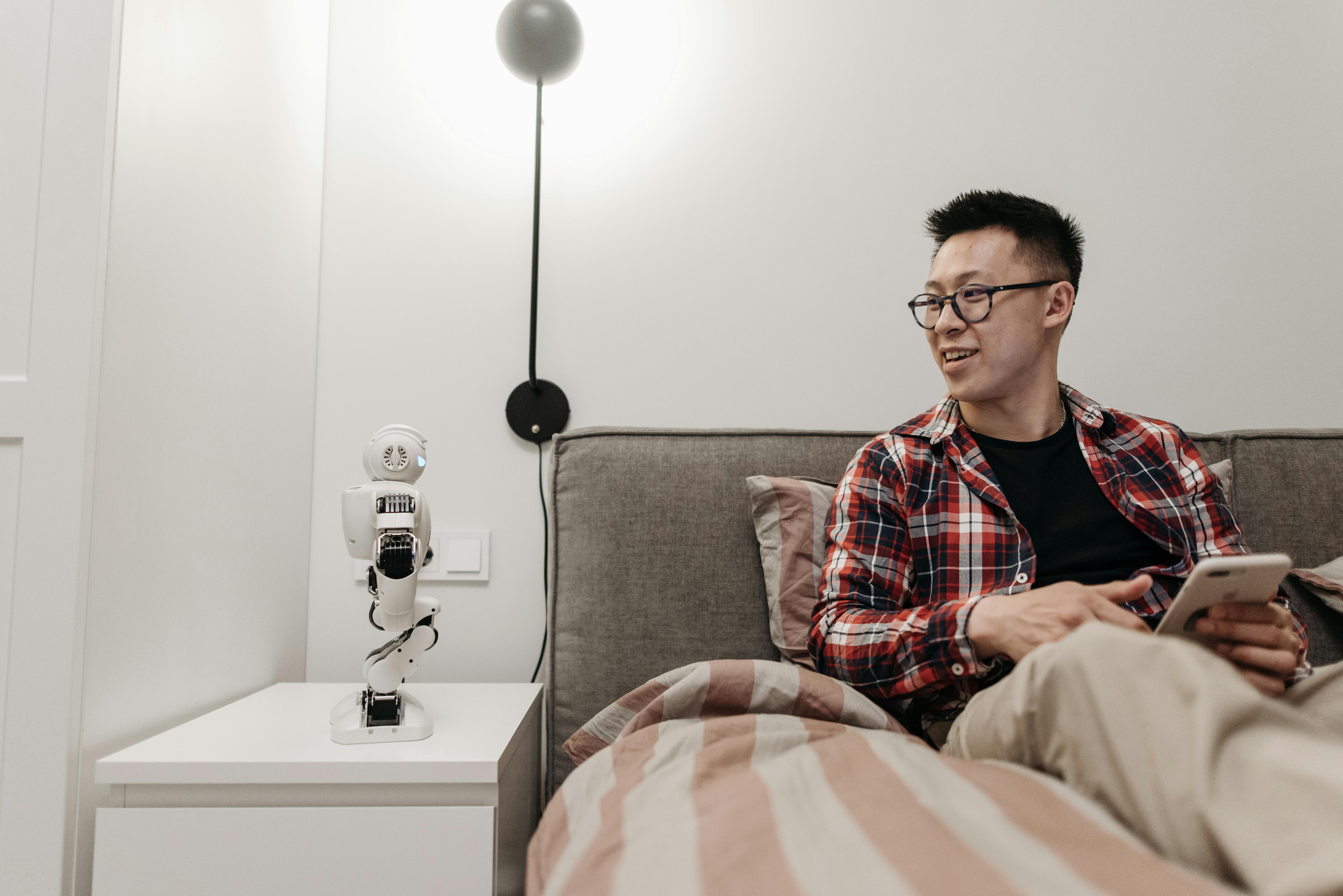 a man in plaid long sleeves sitting on the bed while looking at the robot