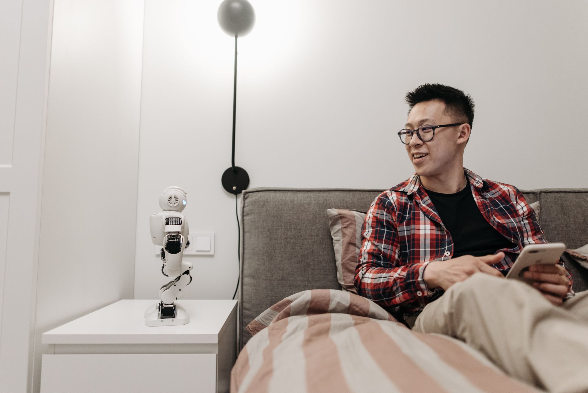 Man sitting on bed using mobile phone, interacting with small robot on bedside table.