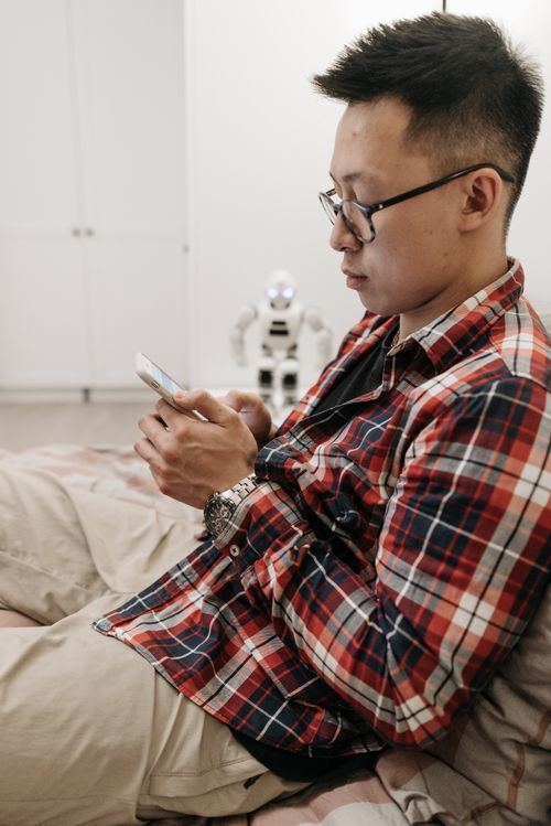 Free Asian man in plaid shirt using smartphone indoors, relaxed atmosphere. Stock Photo