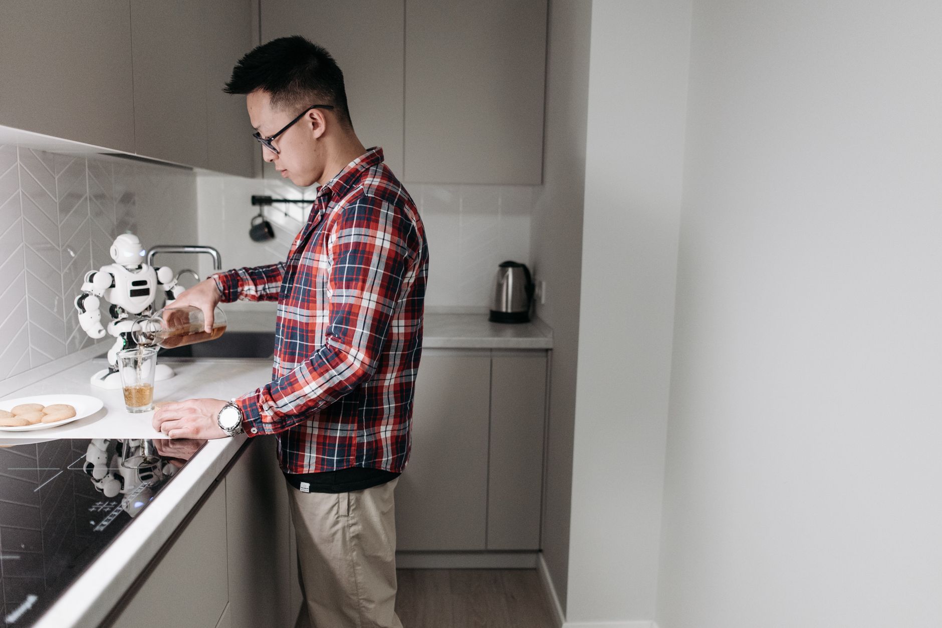 Man in plaid shirt pours drink in a sleek kitchen with a robot.