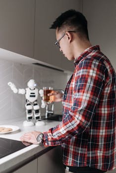 Man in kitchen holding a drink, interacting with a humanoid robot. Modern indoor setting.