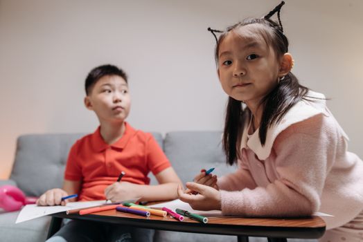 Two Asian children drawing with colored pens indoors, showcasing creativity and sibling bonding.