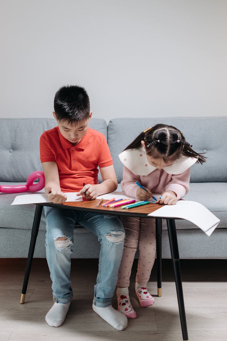 A Girl And Boy Sitting On The Couch While Drawing On Paper Using Colored Pens