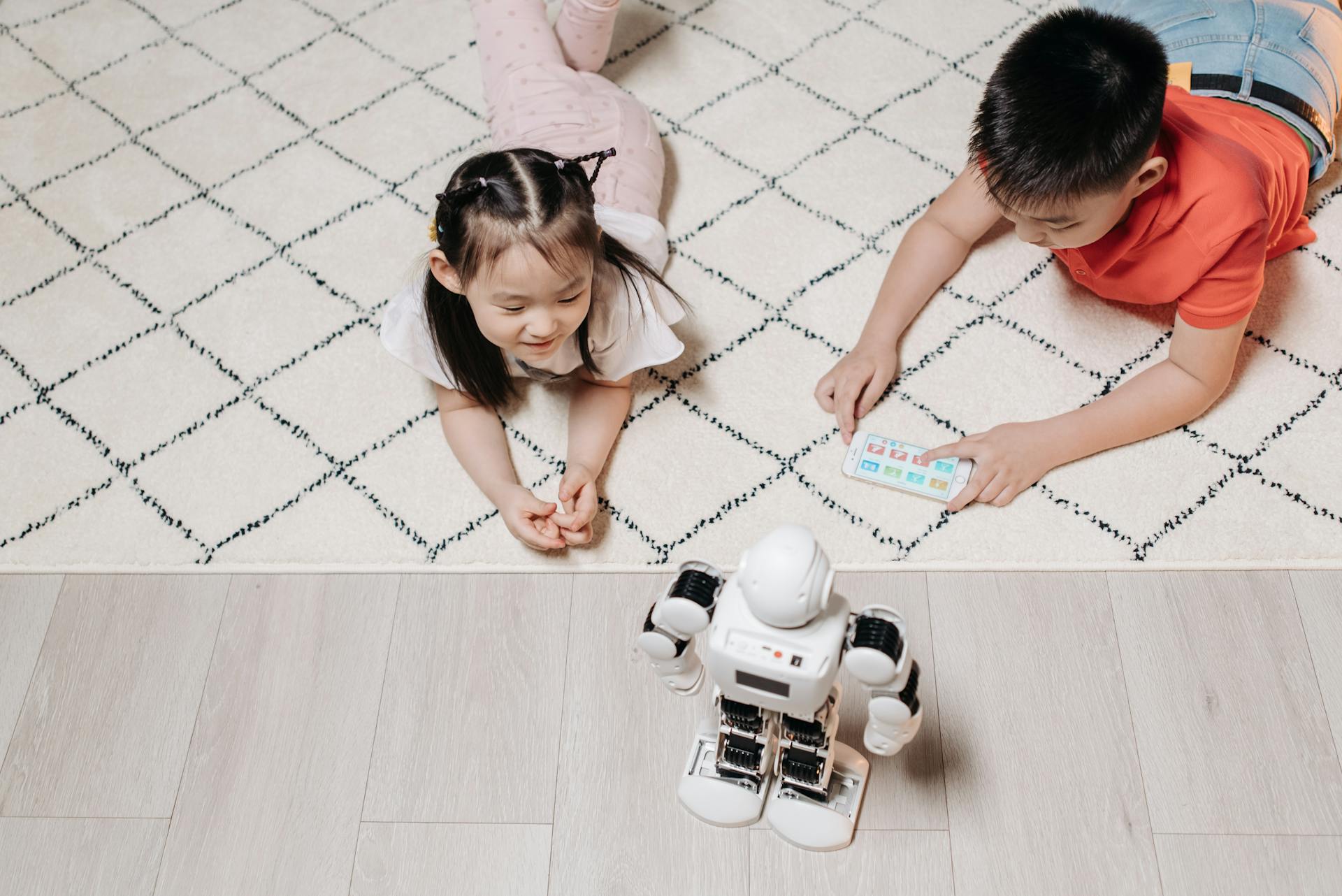 Two children control a playing robot with a mobile device indoors, engaging in technology.