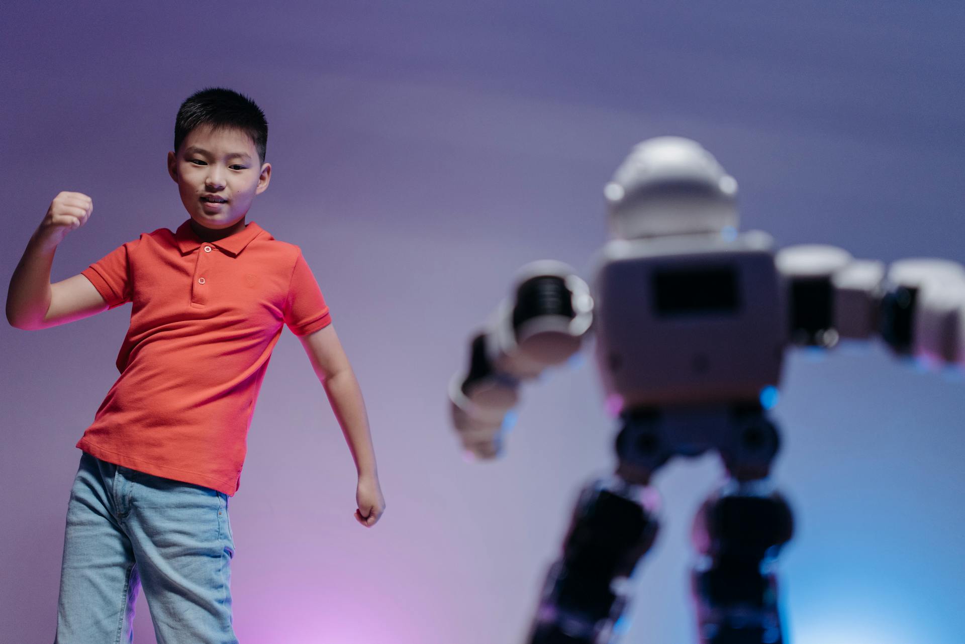 A young boy in a red shirt energetically interacts with a futuristic robot toy under colorful lighting.