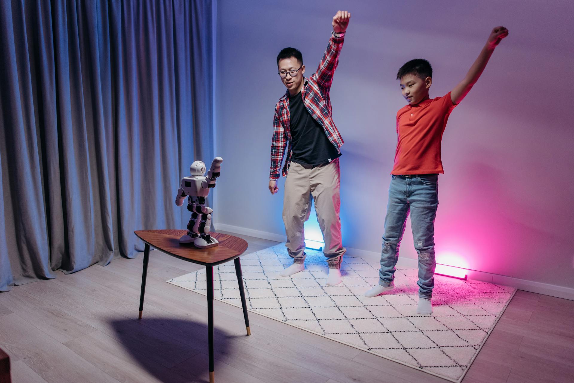 Father and son enjoy dancing with a humanoid robot in a colorful home setting.