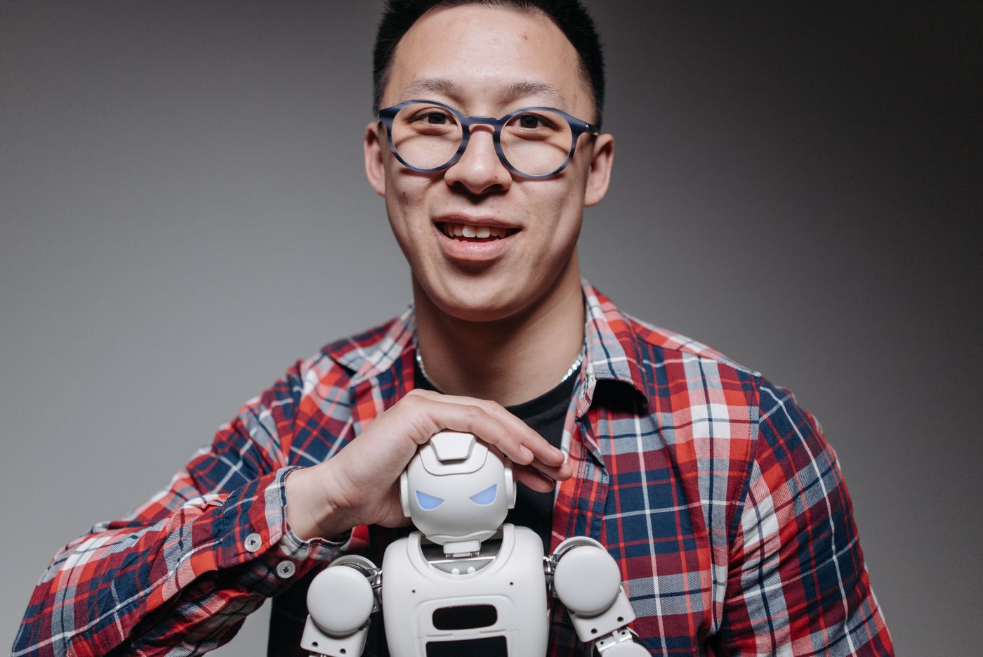 Asian man smiling, holding a small toy robot during a studio shoot. Indoors, with eyeglasses and plaid shirt.