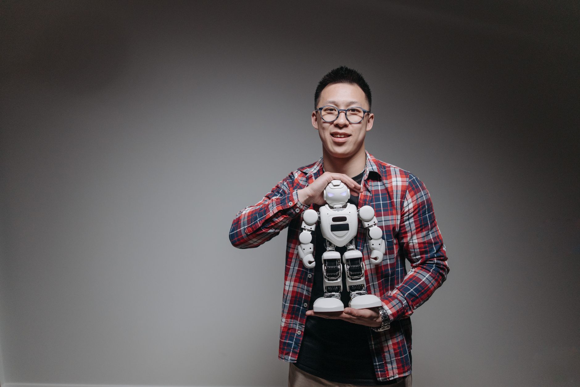 Asian man with eyeglasses holding a toy robot in a studio with a gray background.