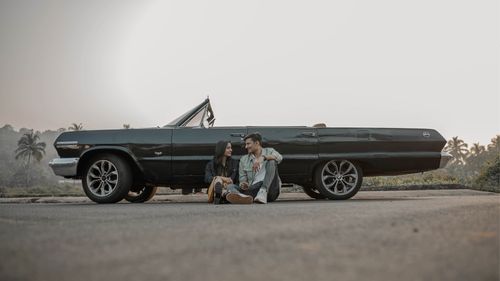 Free A couple sitting on the road beside a vintage convertible in an outdoor setting, conveying a retro vibe. Stock Photo