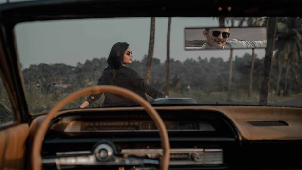 Stylish woman leaning on vintage car as man reflects in rearview mirror, capturing a retro vibe.