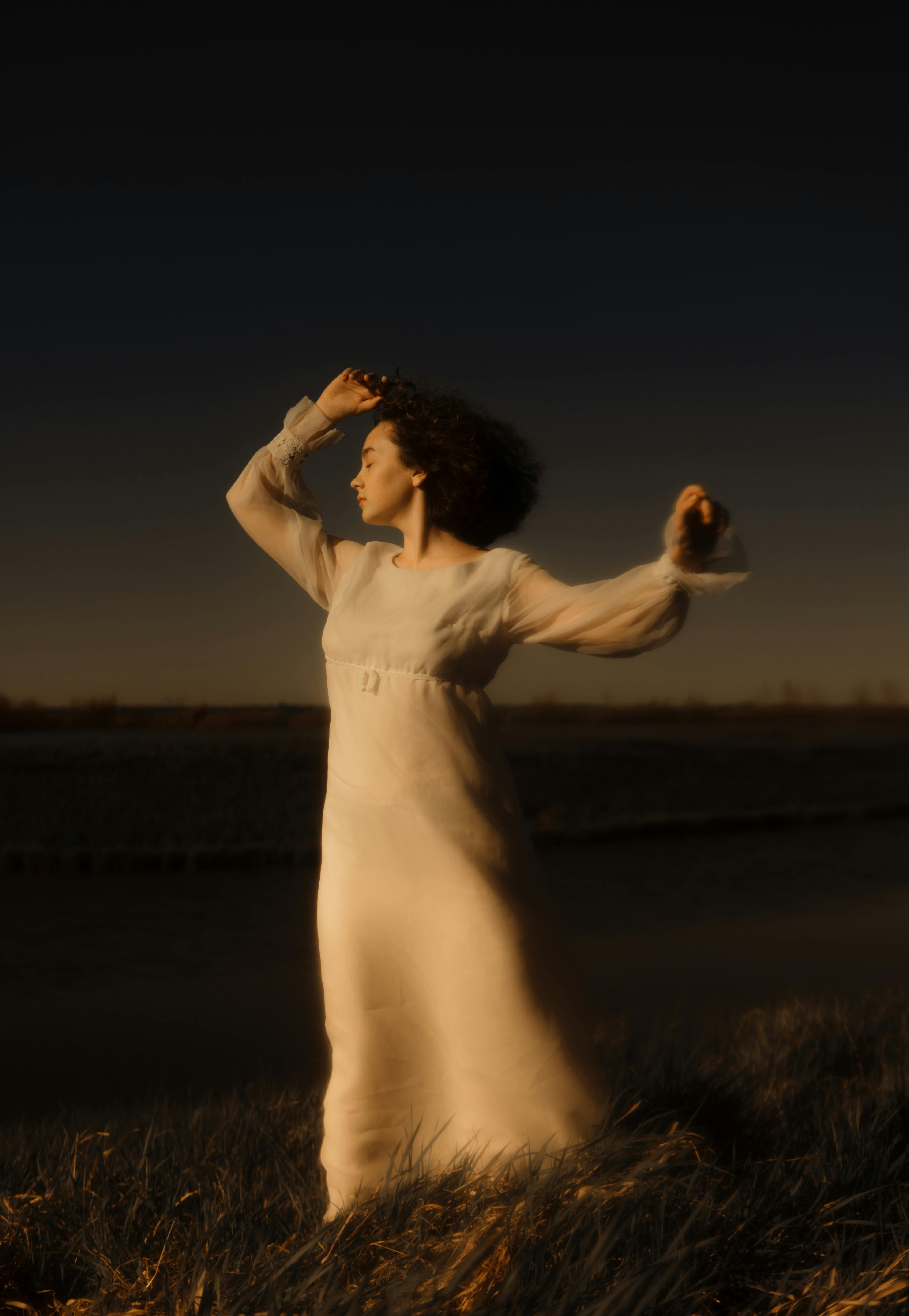 A serene woman in a flowing white dress poses in a grassy field at dusk.