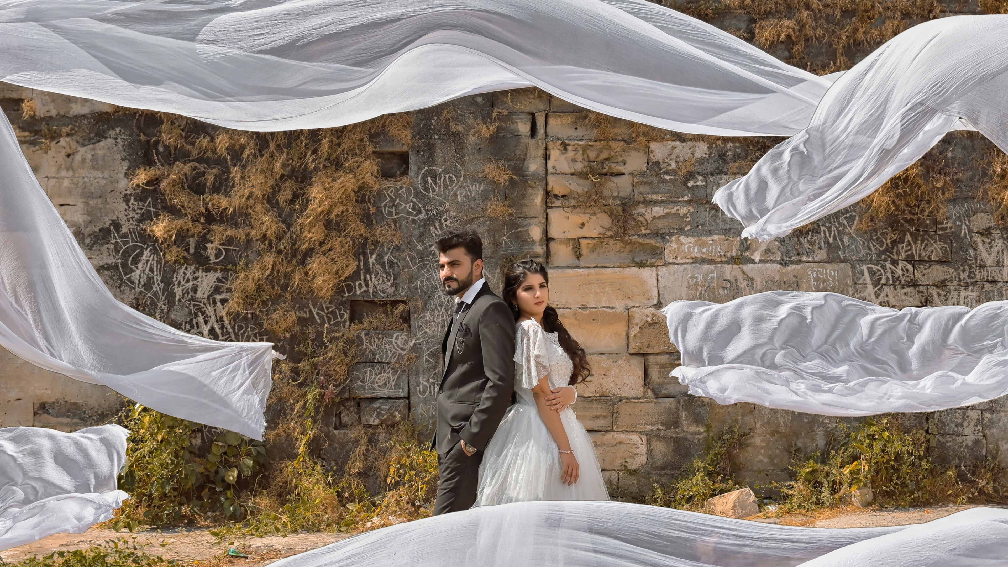 A couple poses elegantly against a rustic brick wall with flowing fabric accentuating the romantic setting.