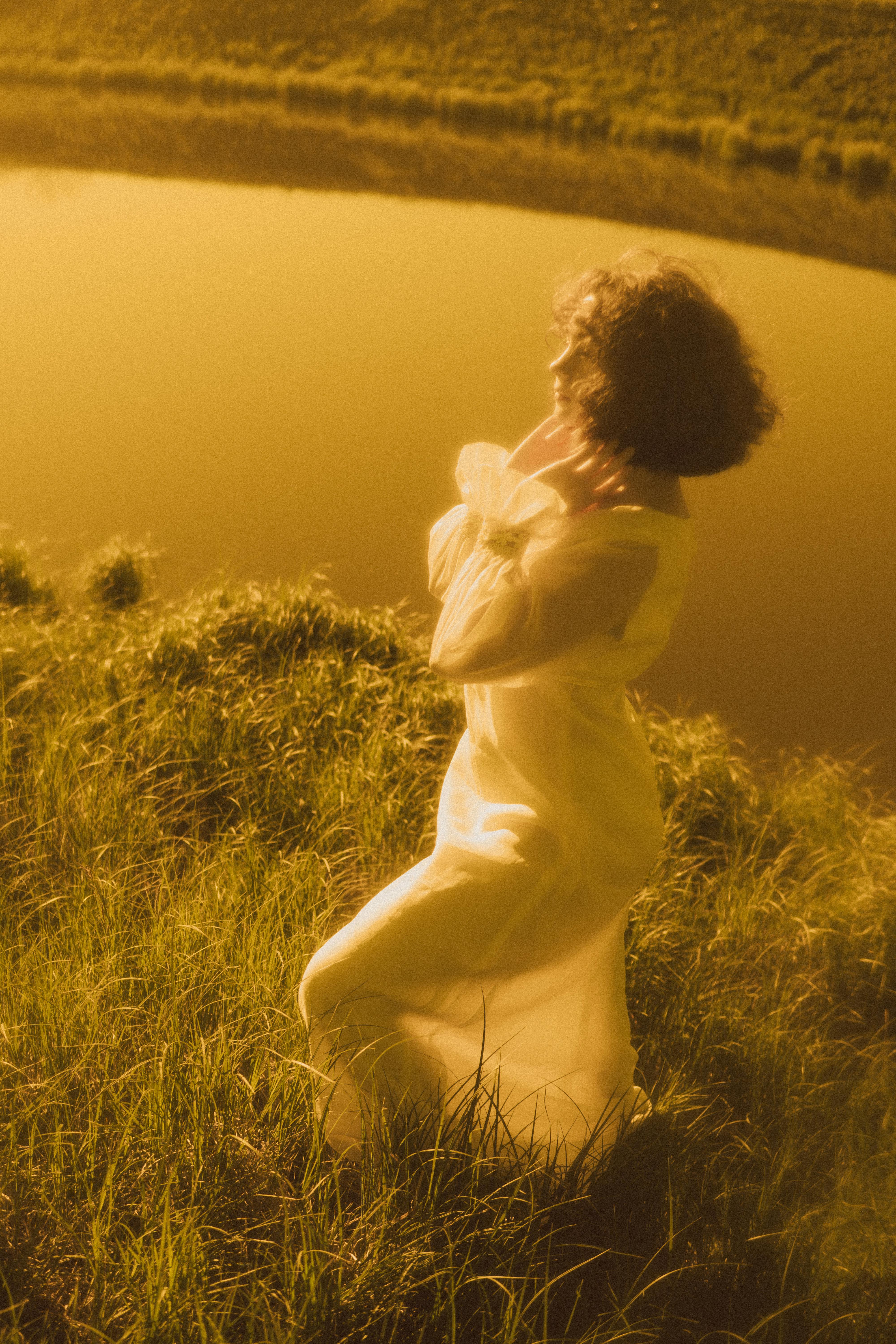 Elegant woman in white dress standing near a lake during golden sunset, exuding tranquility.