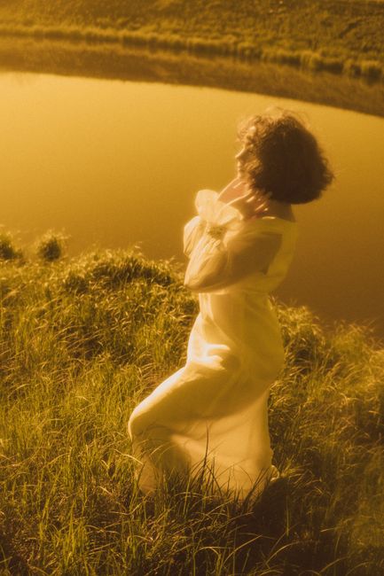 Elegant woman in white dress standing near a lake during golden sunset, exuding tranquility.