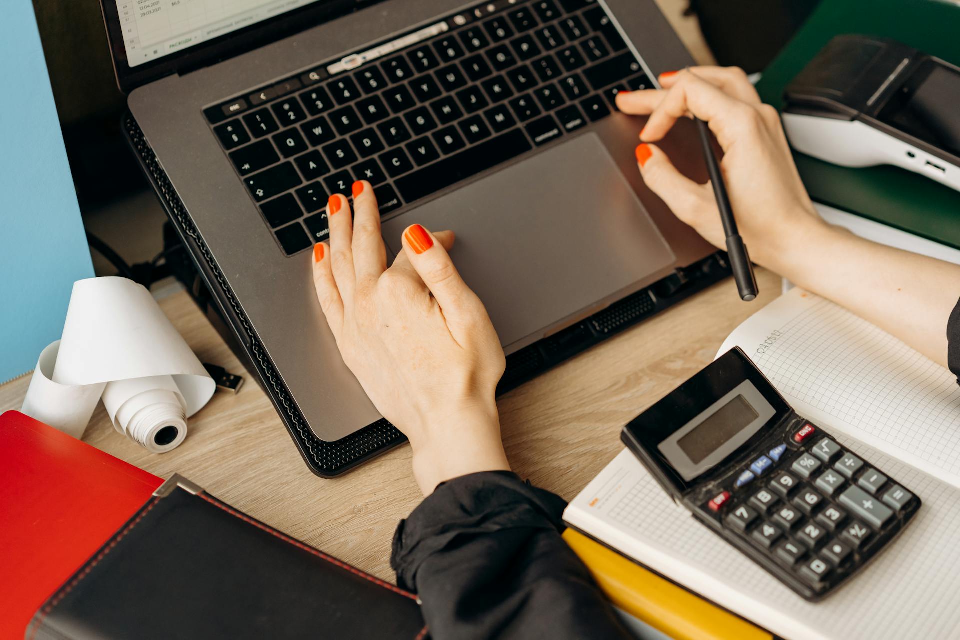 A woman with red nails using a laptop and calculator on a wooden desk.