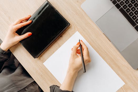 A person writing at a wooden desk with a black notebook and laptop.