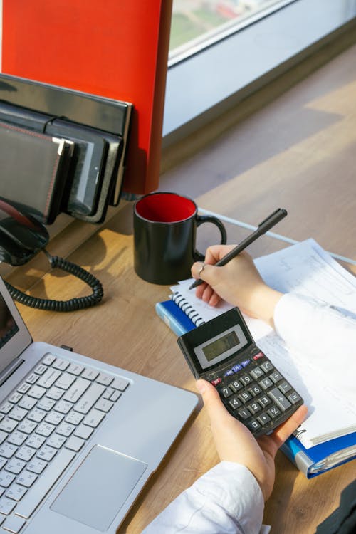 Person Writing on a Notebook while Holding a Calculator