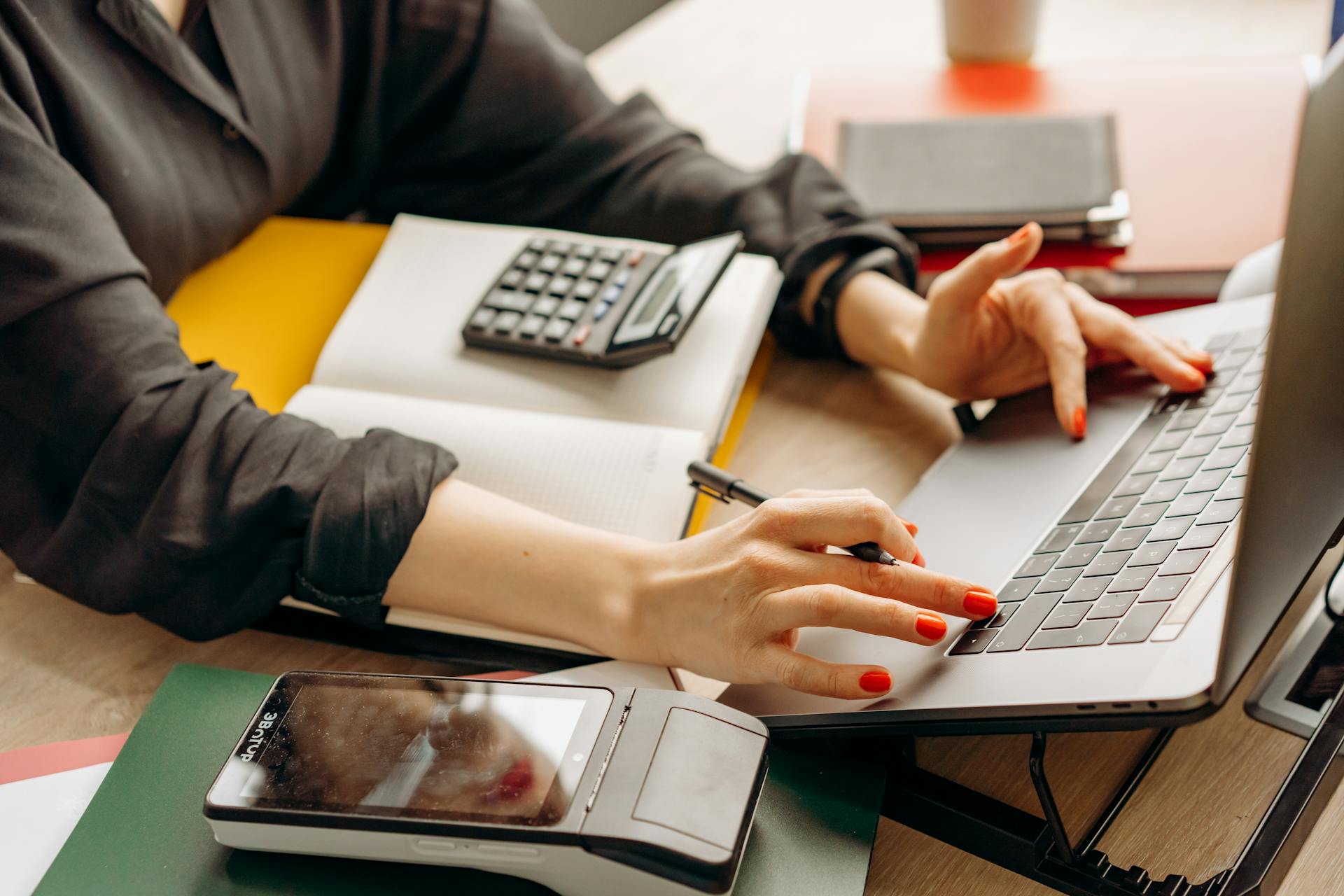 Close-up of a person using a laptop with a payment terminal and calculator on the desk.