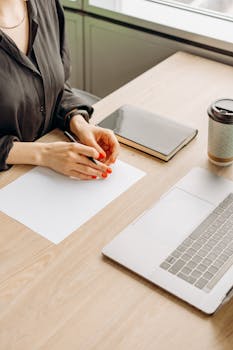 Close-up of a woman's hands with a notebook, coffee, and laptop on a desk.