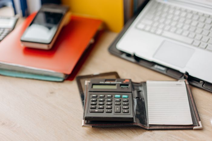 A desk calculator and a notebook beside a laptop on a wooden desk