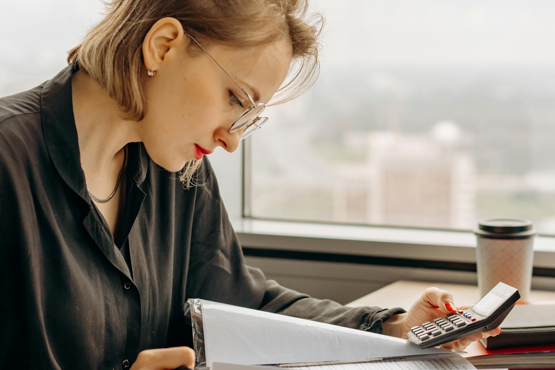 Businesswoman working with calculator and financial documents by window, focused and analytical.