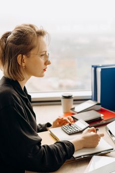 Woman at office desk using a calculator, focused and serious, with a city view.