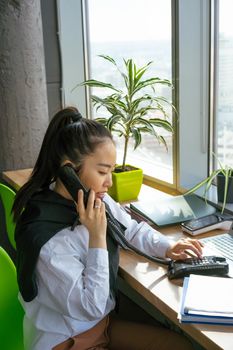 Businesswoman multitasks at her office desk, talking on phone and working on a laptop.