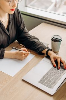 Businesswoman writing notes while working on a laptop at a wooden desk with a coffee cup nearby.