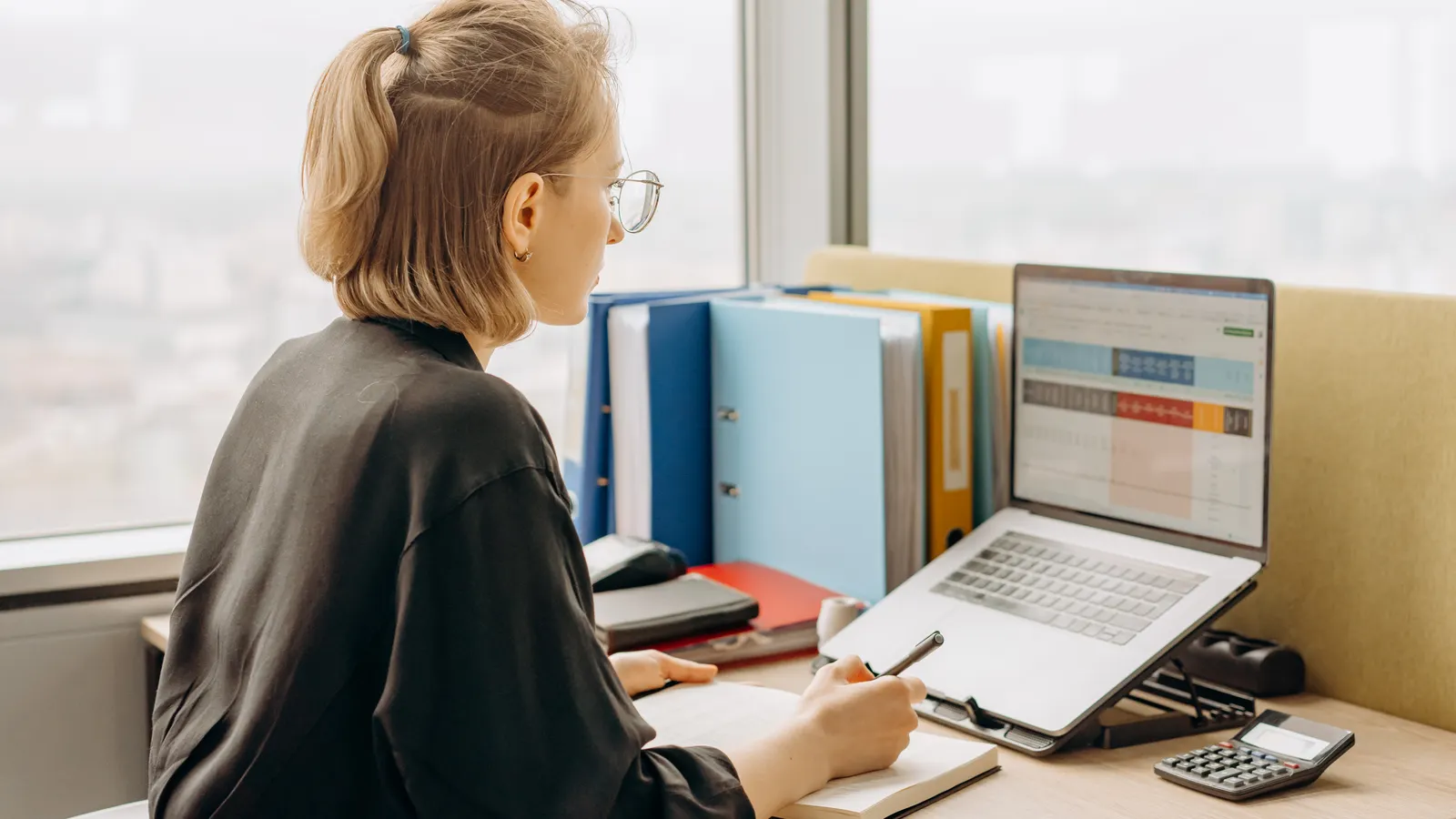 An analyst working through a spreadsheet at a desk, with a notebook open beside the laptop