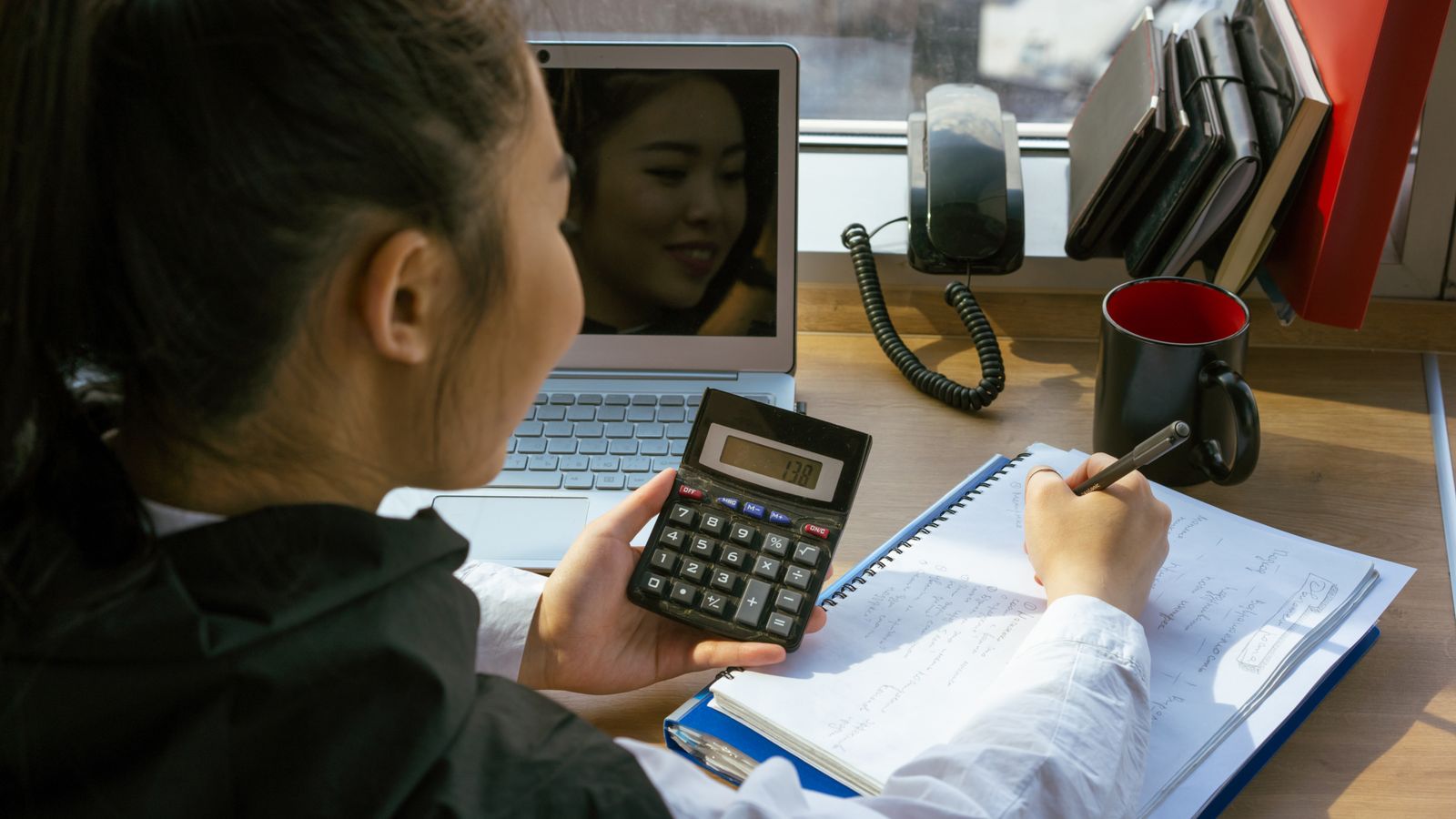 A woman writing in a notepad while using a calculator at a desk with a laptop