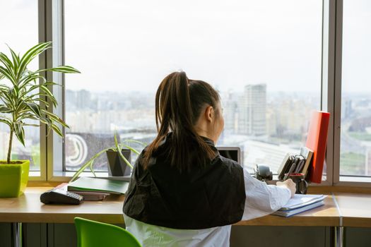 Woman writing at desk with a view of the city skyline through a large window.