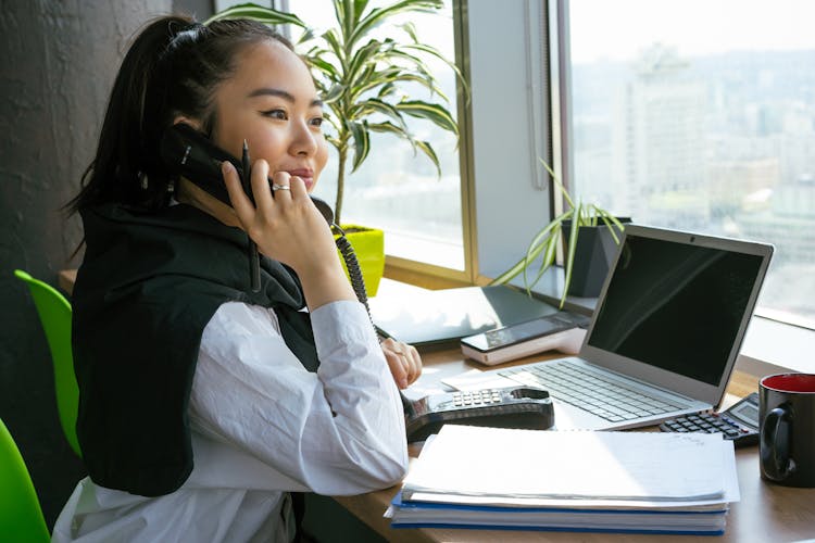 A Woman Sitting On A Chair In Front Of Her Workstation While Having A Telephone Call