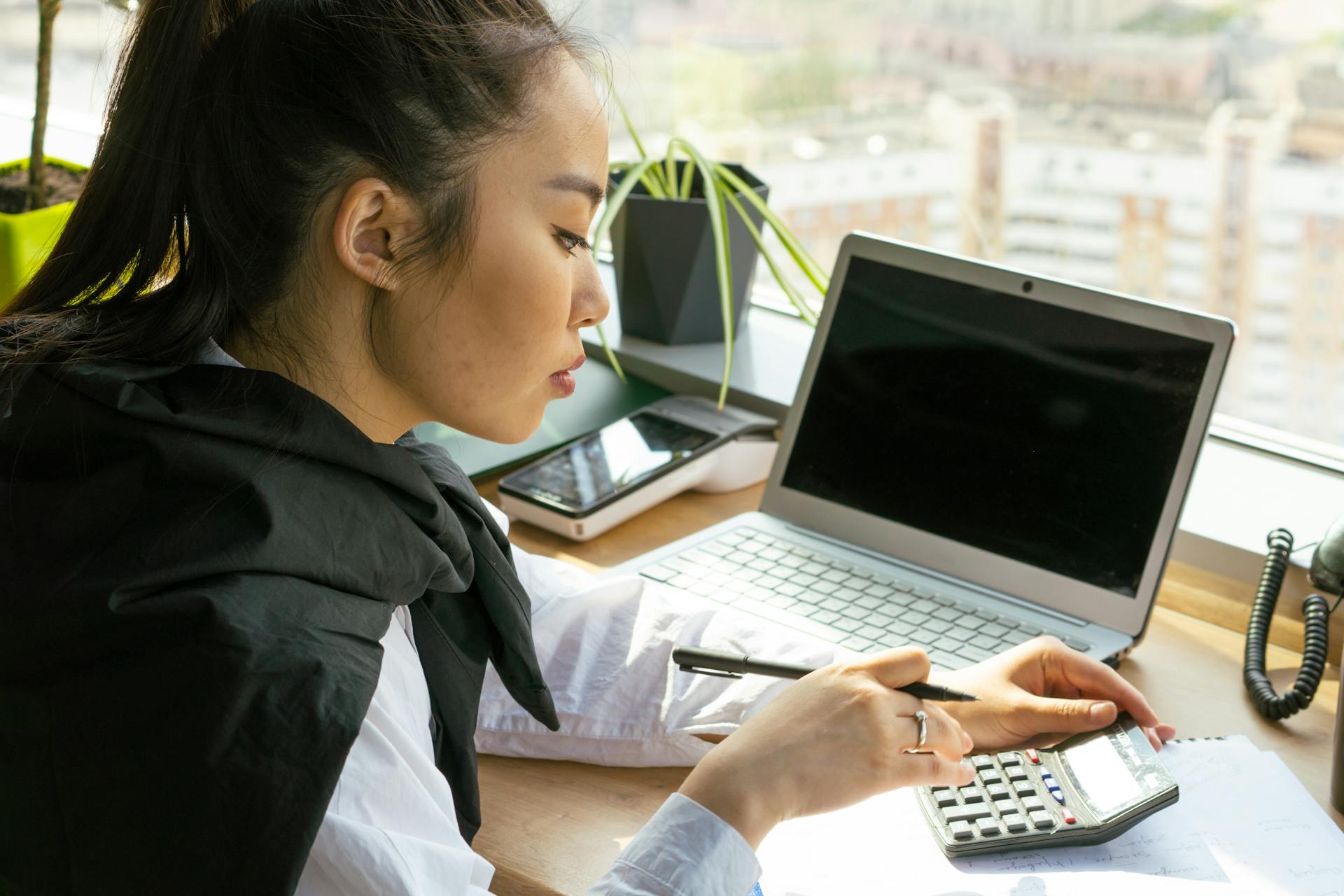 Asian woman working in a bright office, calculating finance on a desk with a laptop.