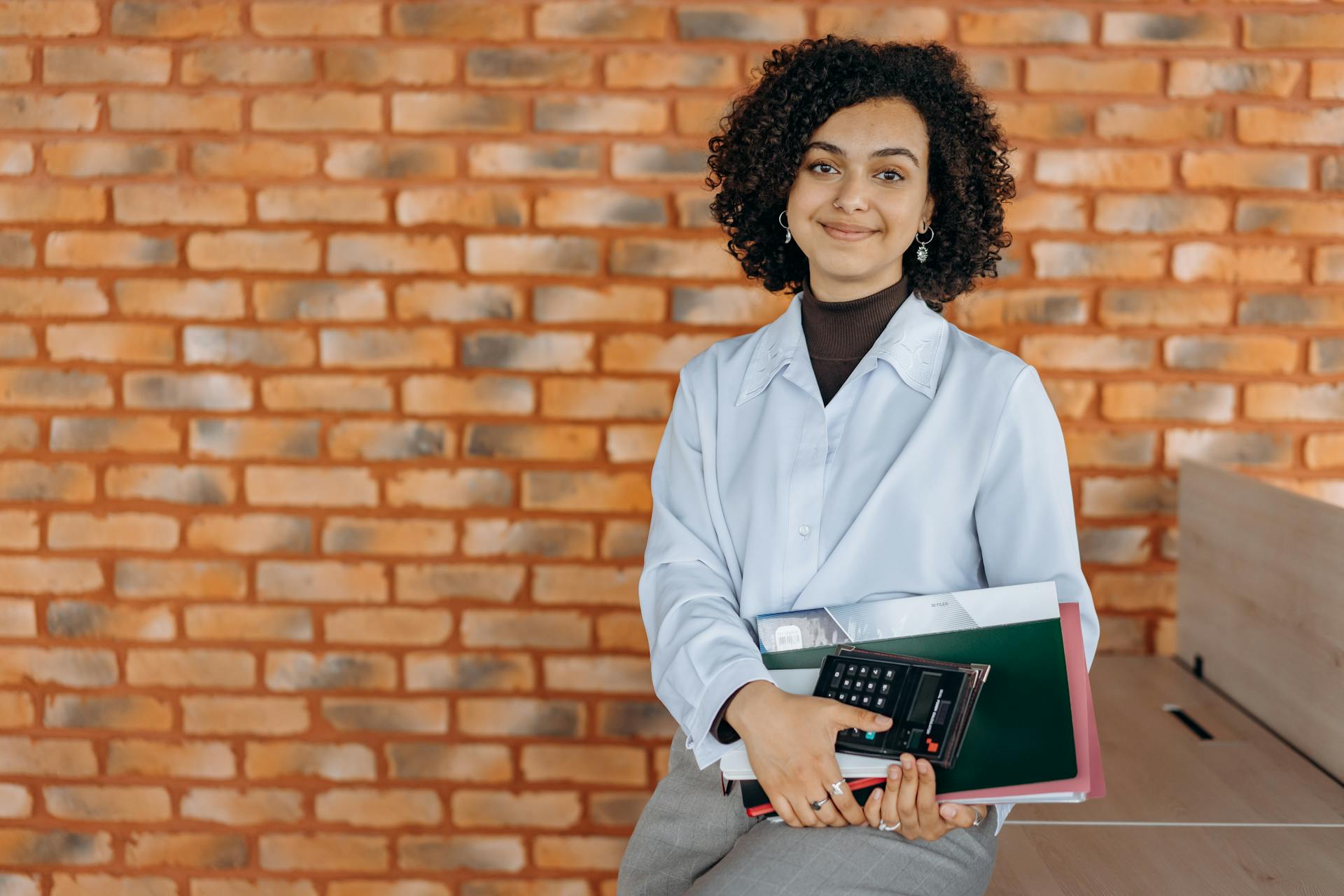 Confident woman holding calculator and files against a brick wall, exuding professionalism and warmth.