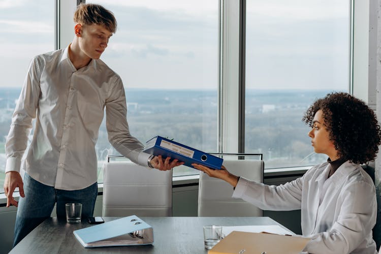 Young Man Standing In White Long Sleeve Shirt Giving A Book Report To His Coworker Inside A Conference Room