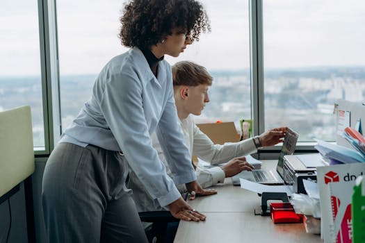 Two colleagues collaborating at a desk in a modern office setting, focused on a laptop.