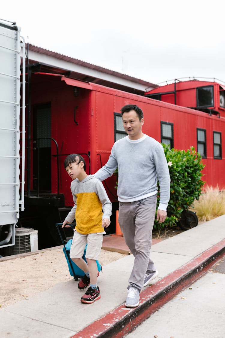 A Man Walking With A Child Pulling A Blue Luggage Near A Red Train