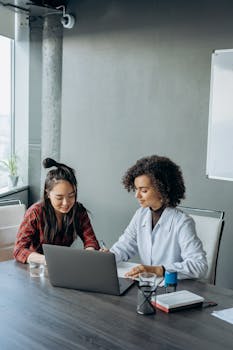 Two women collaborating on a project in a modern office setting with a laptop and documents.