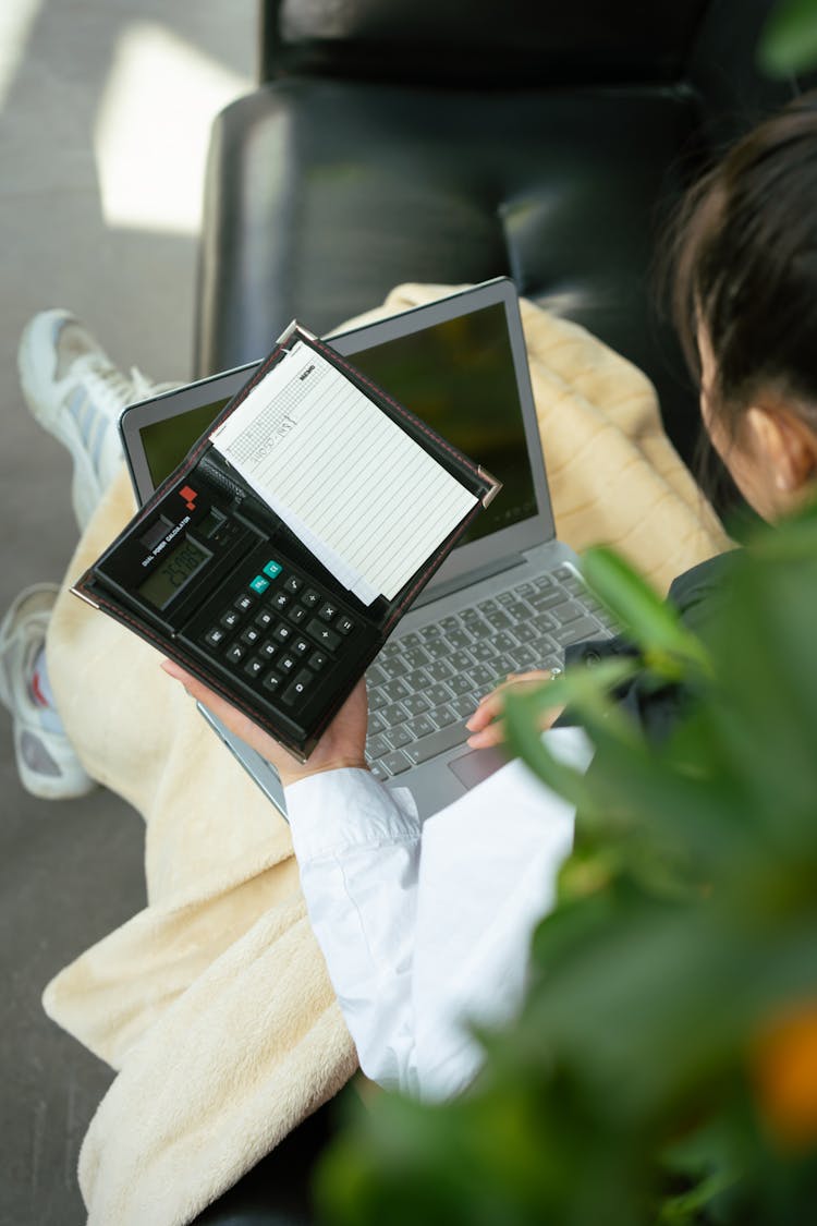 A Person Computing Something On The Calculator While Typing On Her Laptop