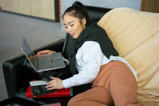Woman using laptop and calculator while relaxing on a couch, embodying remote work ambiance.