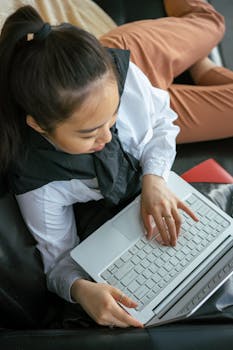 A young woman relaxes on a couch while typing on her laptop, depicting a casual work-from-home setup.