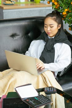 Young woman sitting on a black sofa, using a laptop, with a cozy blanket indoors.