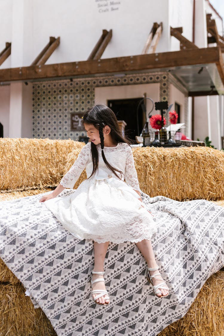 Cute Little Girl In White Dress Sitting On Printed Blanket On A Pile Of Hay