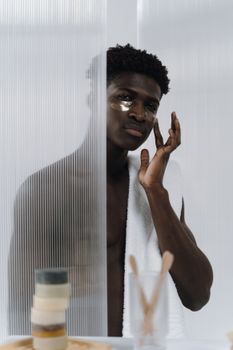 Shirtless man using under eye mask and skincare products in minimalist setting.