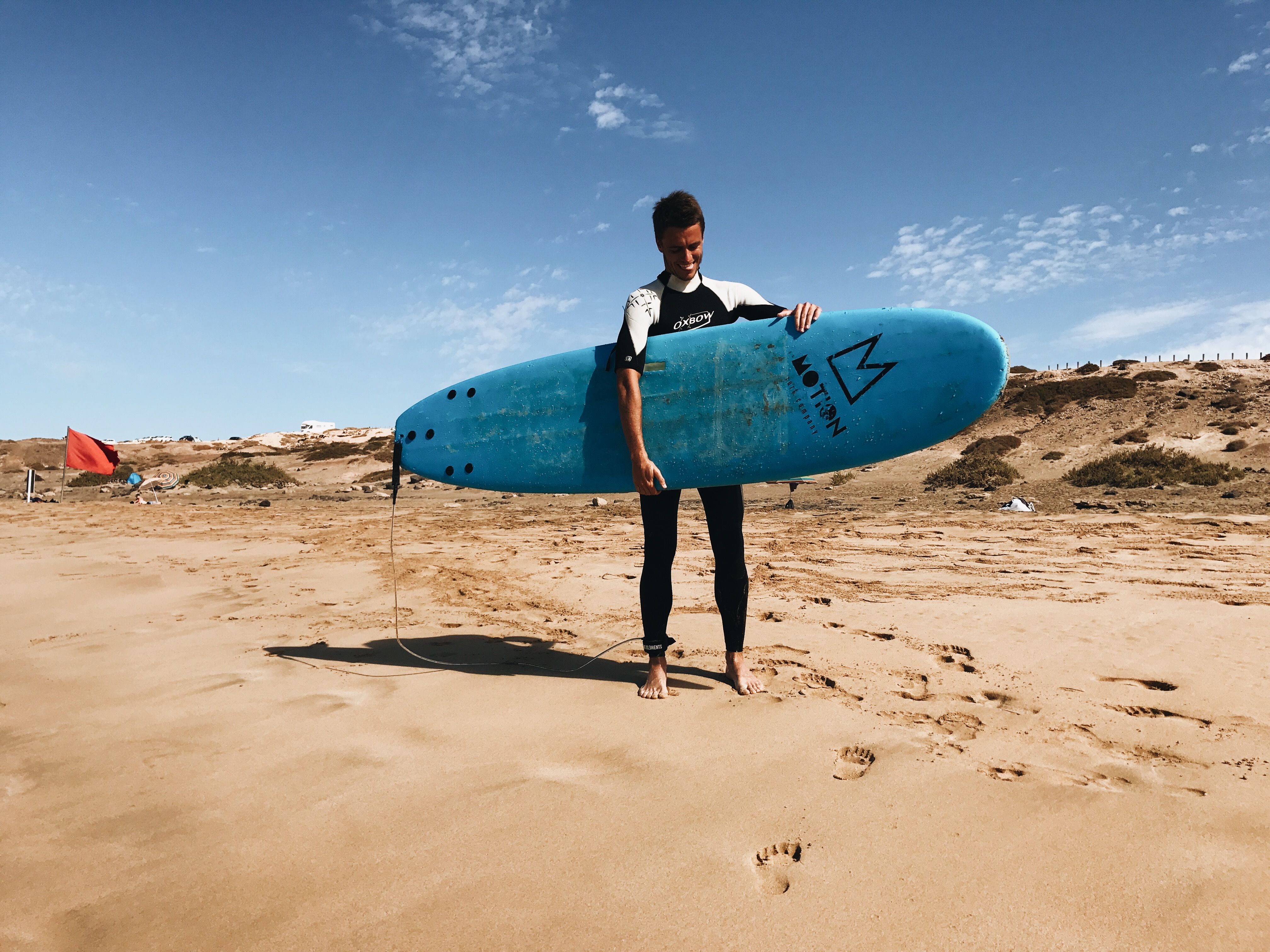 Male surfer holding a blue surfboard on a sunny sandy beach, ready to surf the waves.
