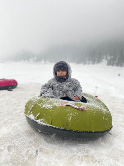 Child in Gray Jacket Riding Green and Black Snow Sled on Snow Covered Ground