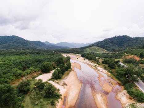 A breathtaking aerial view of a mountain stream surrounded by lush green hills and forests, captured in bright daylight.
