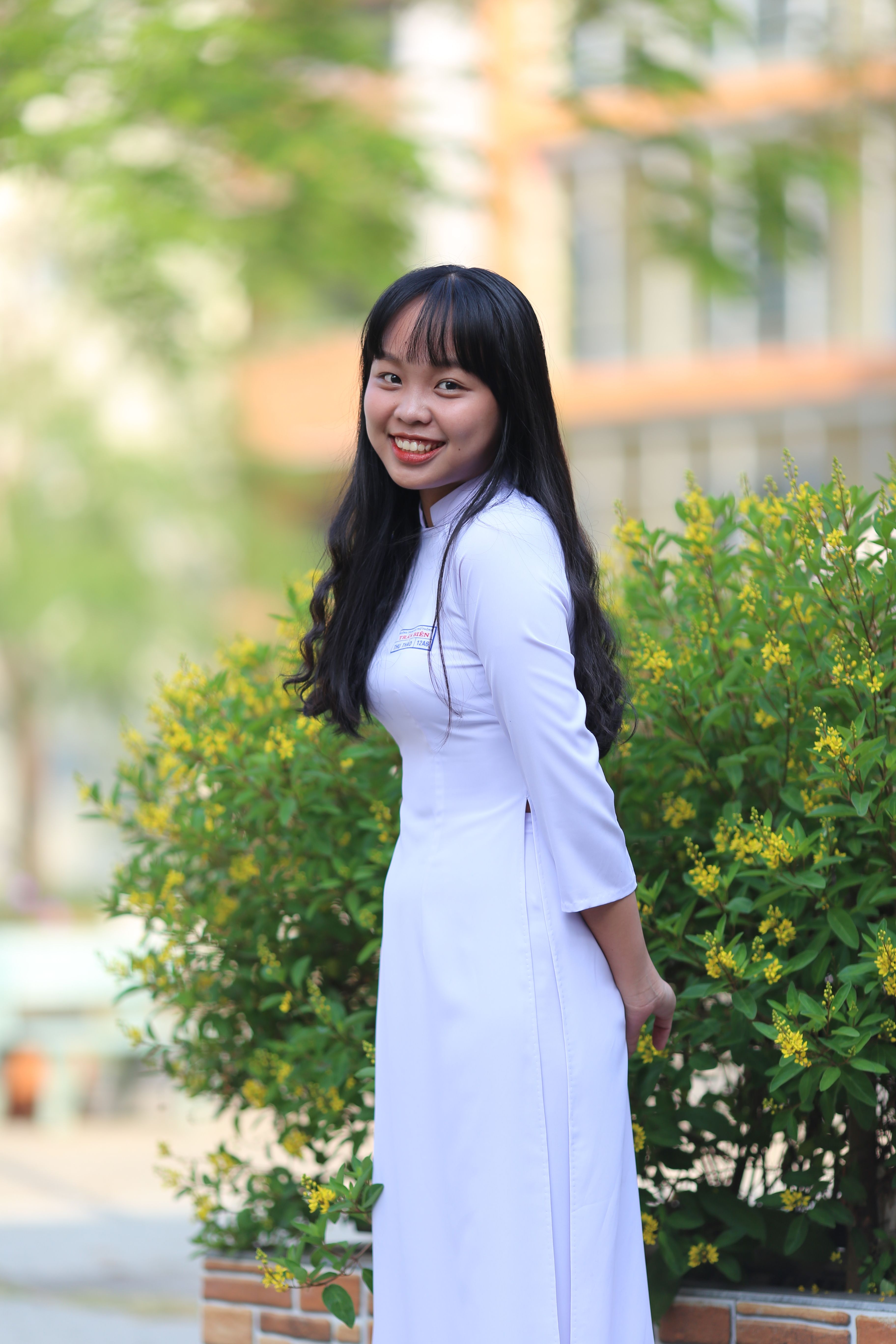 Smiling East Asian woman in a white dress posing outdoors amidst green foliage.